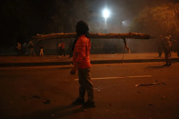 Manifestante con chaqueta naranja y capucha de pie en la calle frente a un tronco atravesado en la vía