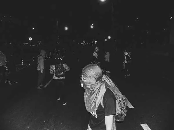 Manifestante con el rostro cubierto por un pañuelo camina de noche en una calle oscura, en blanco y negro