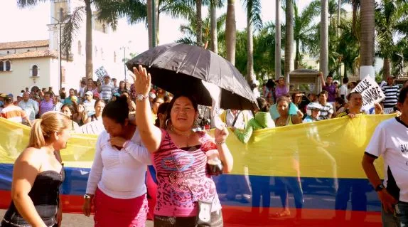 Manifestantes con una bandera de Colombia y un paraguas negro en una marcha bajo las palmeras