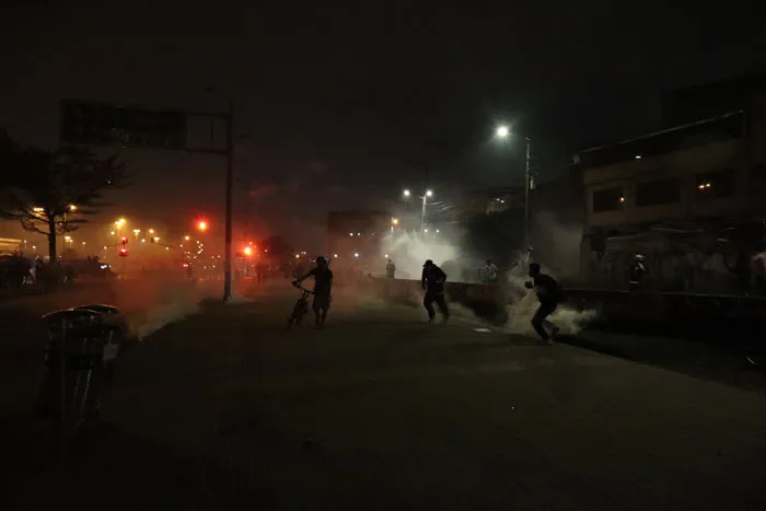 Siluetas de manifestantes entre humo y luces rojas en una calle oscura durante una protesta