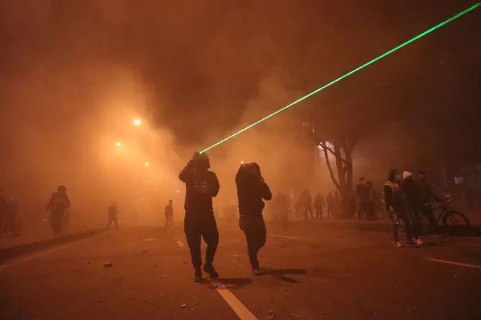 Manifestantes entre humo naranja y un rayo láser verde que cruza la calle
