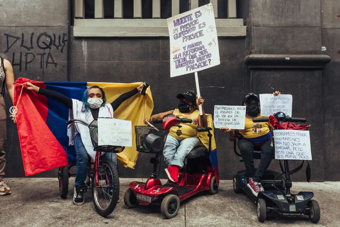 Manifestantes en scooters motorizados y bicicleta con bandera de Colombia y carteles frente a un muro