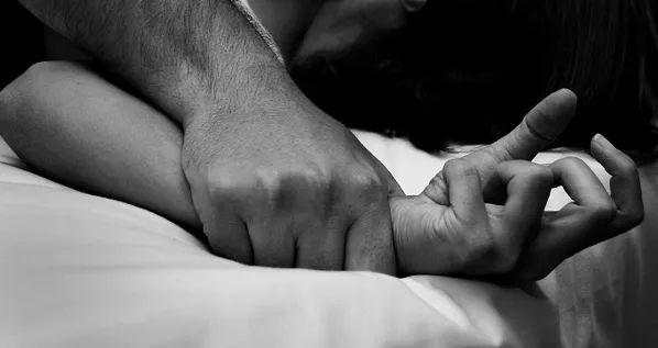 Mano de un hombre sujetando con fuerza la muñeca de una mujer sobre una cama, en blanco y negro
