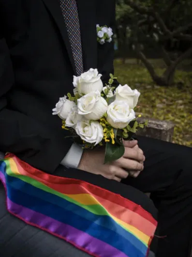 Manos sosteniendo un ramo de rosas blancas y una banda con los colores del arcoíris junto a una persona de traje