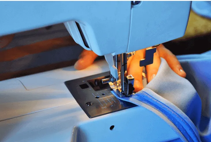 Primer plano de una máquina de coser azul que cose una tela gris con ribete azul