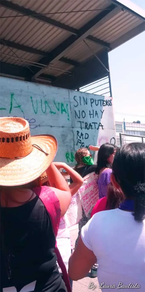 Mujeres con sombrero y pañuelos junto a una pared con la pinta «Sin putero no hay trata» en la marcha del 8M