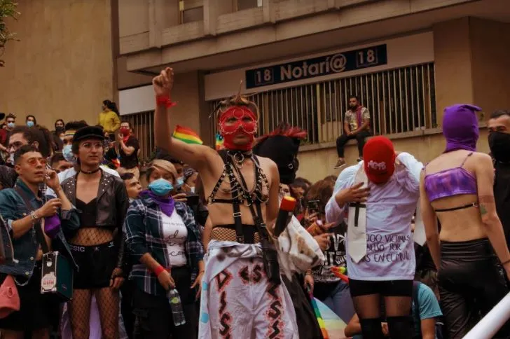 Manifestantes con máscaras rojas y moradas y atuendos de cuero frente a una notaría, entre la multitud del Orgullo