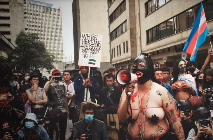Manifestante con megáfono y máscara negra, rodeado de una multitud con banderas trans y un cartel, en la Marcha del Orgullo
