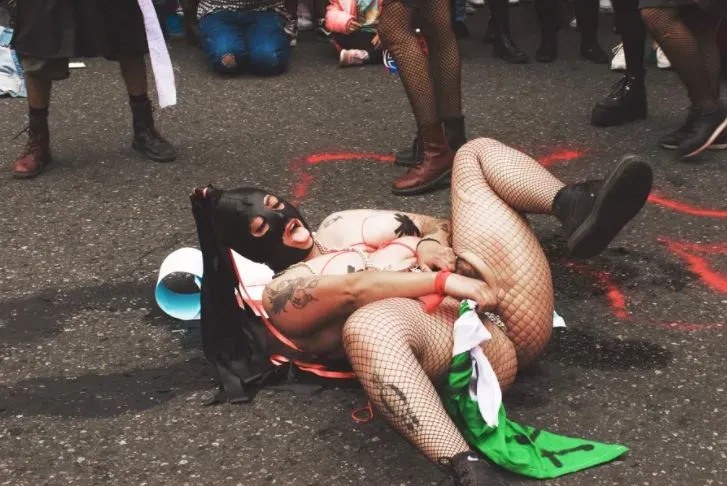 Performer enmascarado con medias de malla tendido en el asfalto junto a marcas rojas, ante la multitud