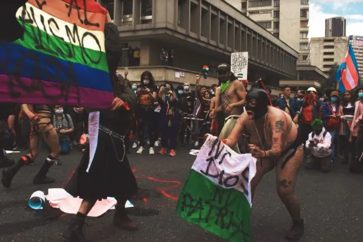 Performance en la calle con personas enmascaradas, una bandera arcoíris y una bandera con el mensaje «Ni Dios ni patria»