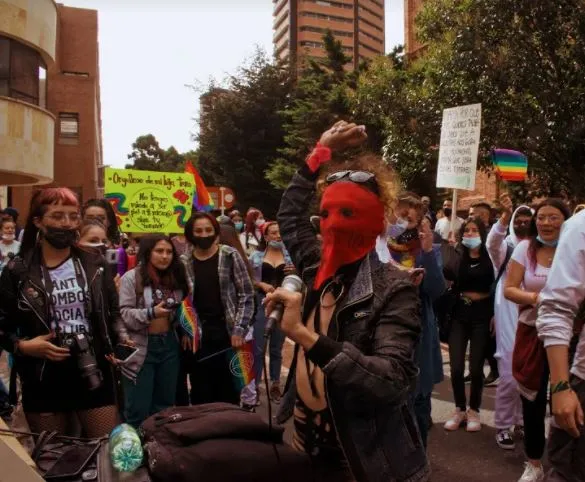 Persona con máscara roja y chaqueta negra habla entre la multitud, con carteles y banderas arcoíris al fondo