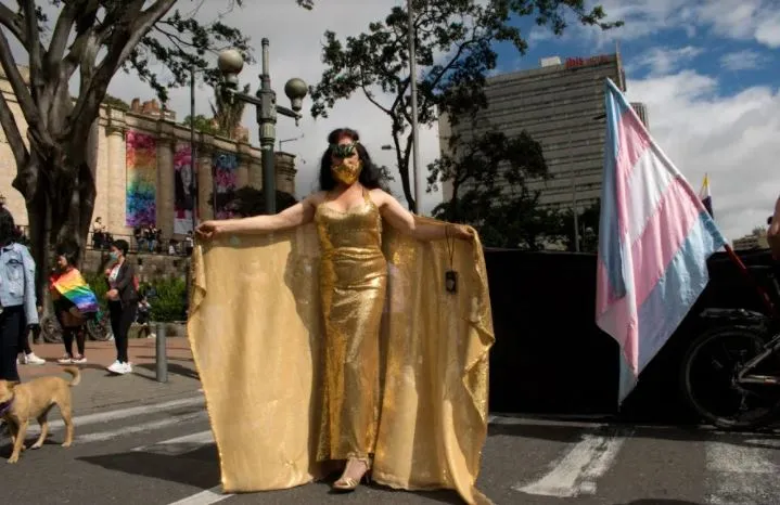 Persona con vestido dorado largo y capa posa en la calle junto a una bandera trans en la Marcha del Orgullo