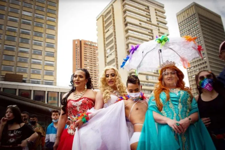 Grupo con vestidos de gala y una sombrilla blanca decorada posa en la Marcha del Orgullo frente a edificios altos