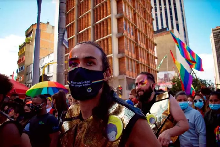 Asistentes con mascarilla y atuendos brillantes ante edificios altos y banderas arcoíris en la Marcha del Orgullo