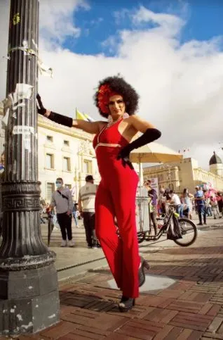 Persona con mono rojo, guantes negros y peluca posa junto a un poste en una calle de Bogotá