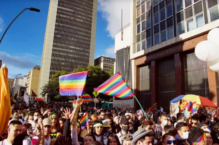 Multitud con banderas arcoíris en una plaza entre edificios altos bajo un cielo azul en la Marcha del Orgullo