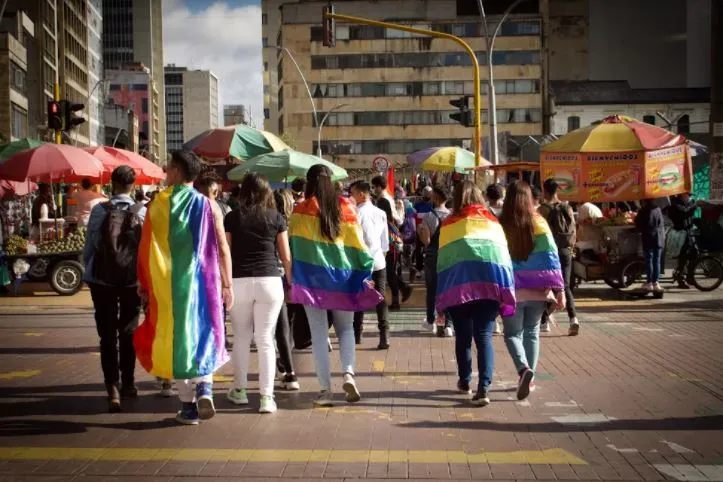 Personas de espaldas con banderas arcoíris sobre los hombros caminan por una calle de Bogotá