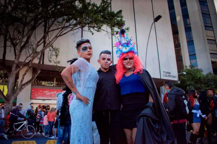 Tres personas con atuendos elaborados, entre ellos un vestido azul claro y un tocado de plumas, posan durante la Marcha del Orgullo