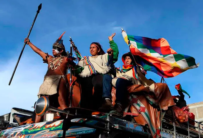 Indígenas de la minga sobre una chiva con lanza y bandera wiphala bajo un cielo azul