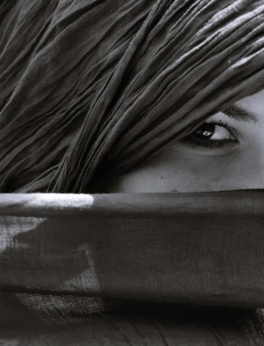 Mirada de una persona a través de mechones de cabello sobre el rostro, con una tela que cubre su boca, en blanco y negro