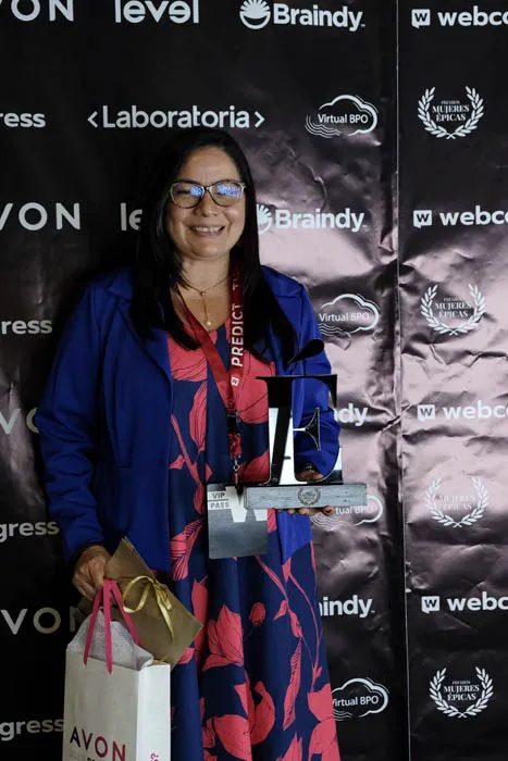 Mujer con abrigo azul sostiene un trofeo y una bolsa de regalo frente al panel de patrocinadores de Mujeres Épicas