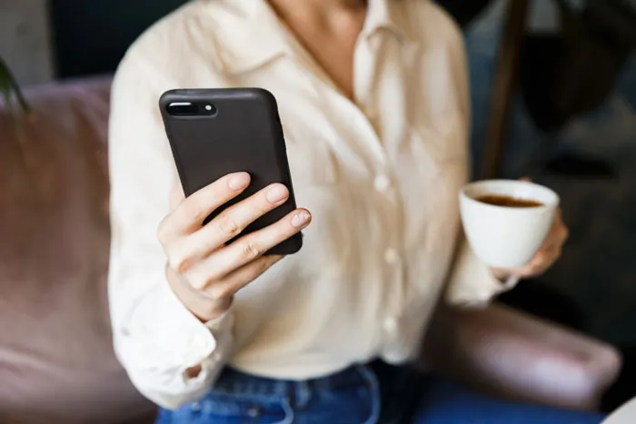 Mujer con blusa clara sostiene un teléfono celular en una mano y una taza de café en la otra