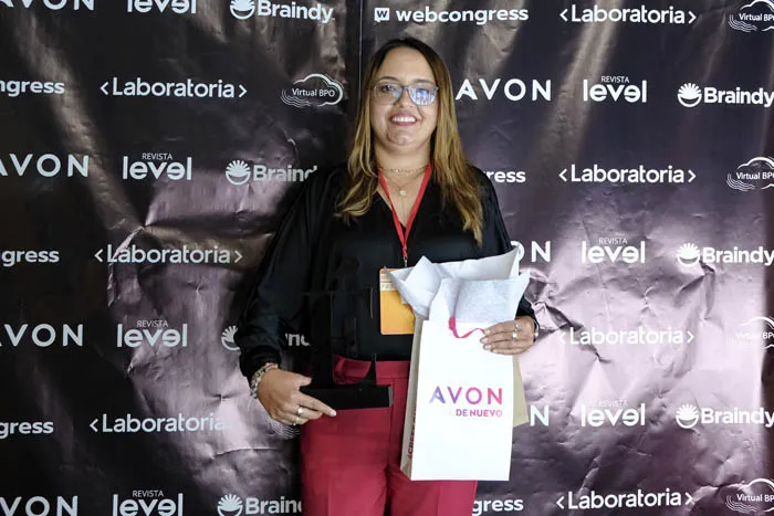 Mujer de blusa negra y pantalón rojo sostiene una bolsa de regalo frente al panel de Revista Level, Braindy y Avon
