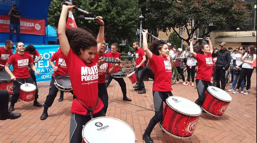 Mujer con camiseta roja levanta los brazos mientras otras tocan tambores rojos en una plaza
