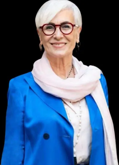 Mujer de cabello blanco con gafas rojas, saco azul y pañuelo claro, sonriendo a la cámara
