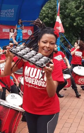 Mujer sonriente con camiseta roja «Mujer poderosa» toca un instrumento de percusión metálico en la ciclovía
