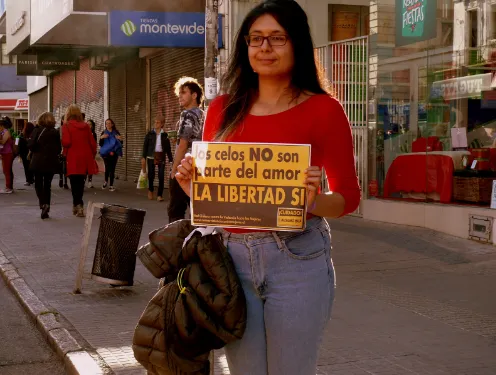 Mujer con gafas y blusa roja sosteniendo un cartel «Los celos NO son parte del amor. La libertad SÍ» en una calle de Montevideo