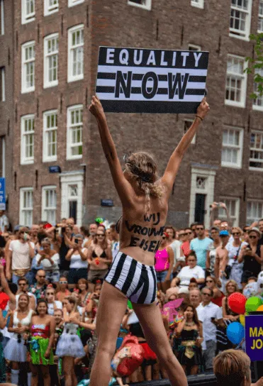 Mujer de espaldas con los brazos en alto frente a una multitud, bajo un cartel «Equality Now»