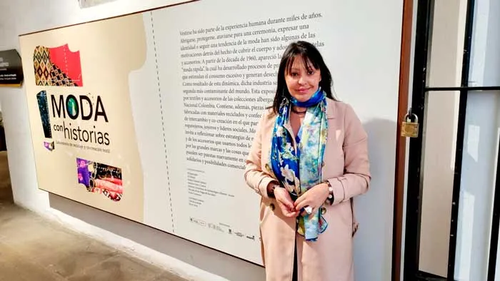 Mujer con chal azul estampado y abrigo rosado junto al panel de la exposición «Moda con historias»
