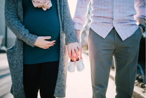 Mujer embarazada con las manos sobre el vientre junto a un hombre que sostiene unos zapatitos de bebé
