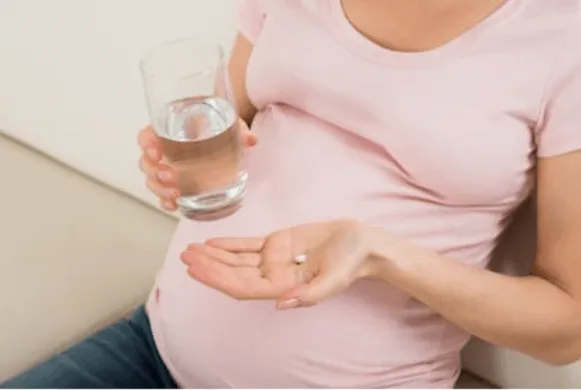Mujer embarazada con blusa rosa sostiene un vaso de agua y una pastilla en la mano