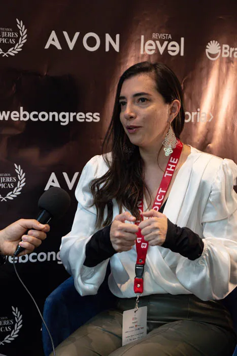Mujer con blazer blanco es entrevistada con un micrófono frente al panel de Revista Level y Avon