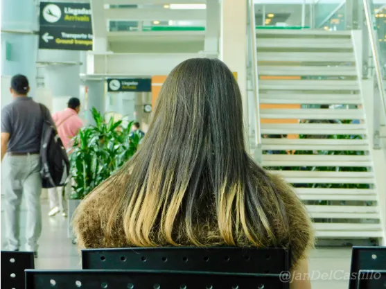 Mujer de espaldas con el cabello con mechas rubias y chaleco de piel, sentada en un aeropuerto