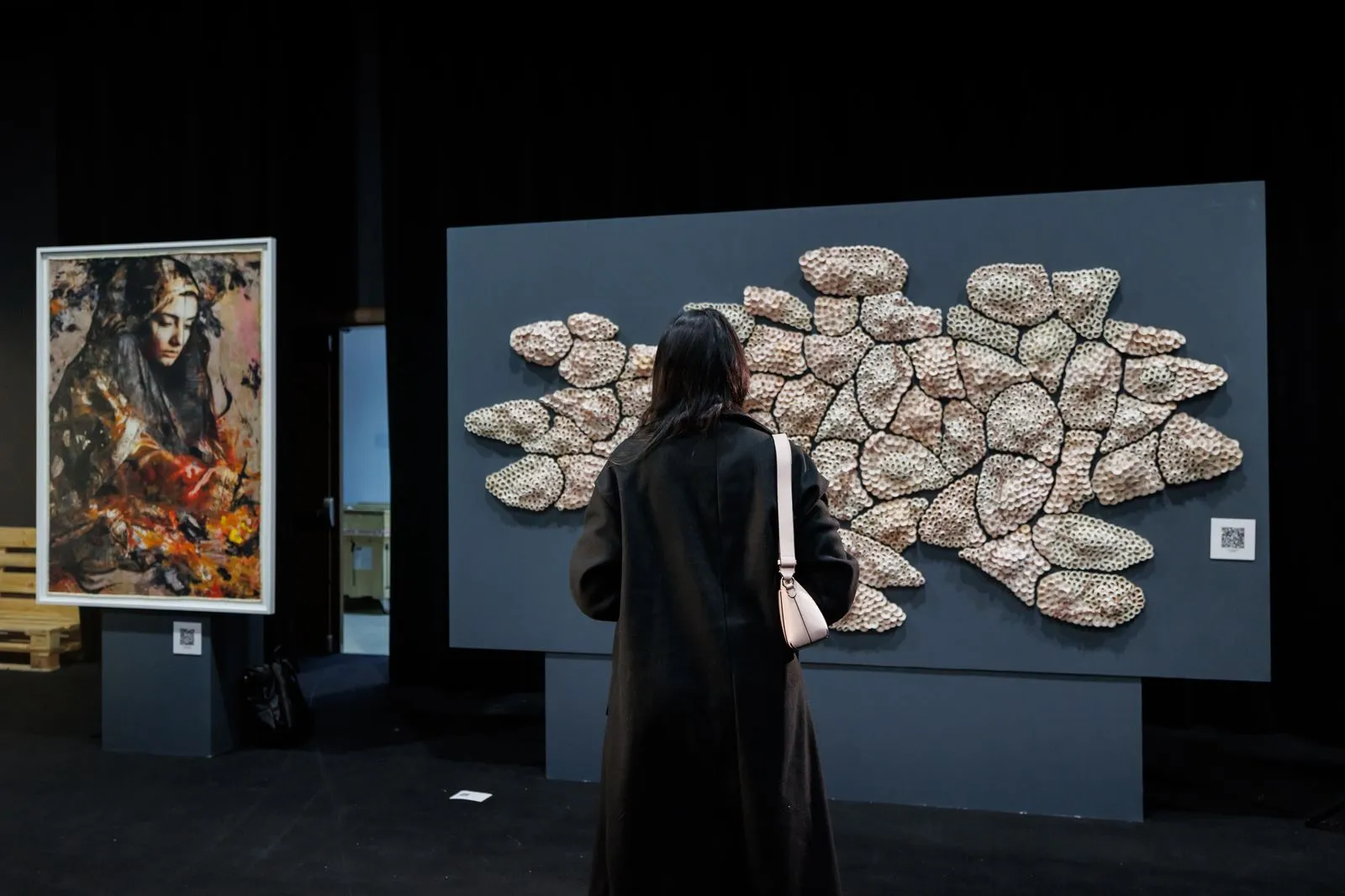 Mujer de espaldas frente a una escultura de piedras en un panel gris y una pintura