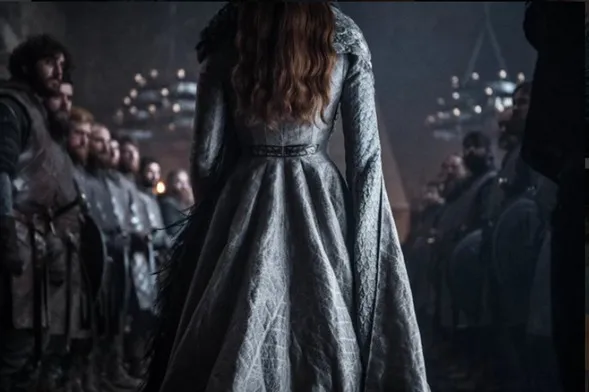 Mujer de espaldas con vestido gris largo frente a una sala con hombres sentados, escena de «Juego de Tronos»