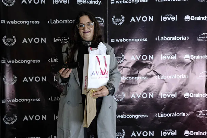 Mujer de abrigo gris con gafas sostiene un trofeo y una bolsa de Avon frente al panel de patrocinadores