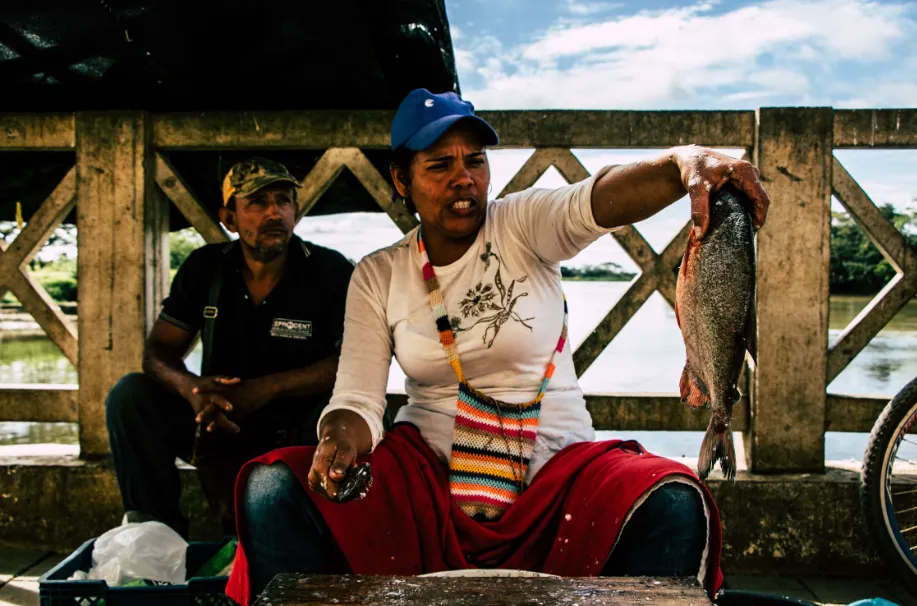 Mujer con gorra azul y falda roja muestra un pescado sentada junto a un hombre en un puente de madera sobre un río