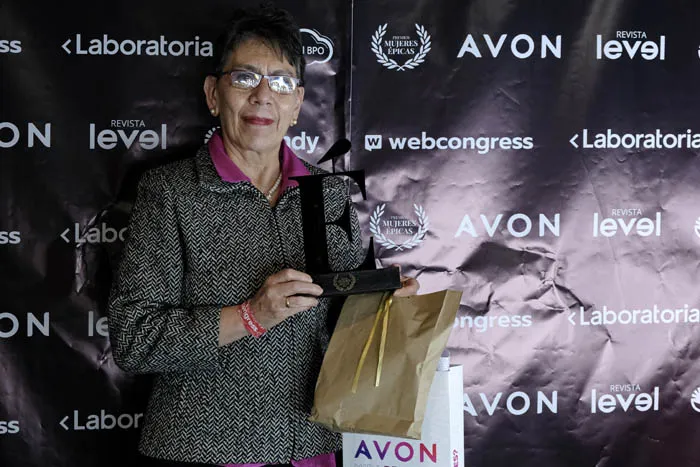 Mujer mayor con gafas sostiene un trofeo y un regalo frente al panel de Revista Level y Avon