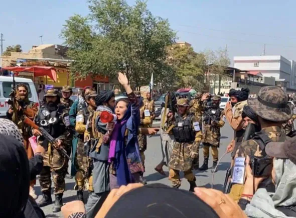 Mujer con megáfono y pañuelo morado frente a hombres armados con uniforme de camuflaje durante una protesta en Kabul