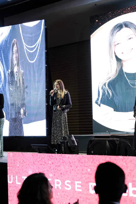 Mujer de pie con micrófono en el escenario ante dos pantallas grandes, con público en primer plano