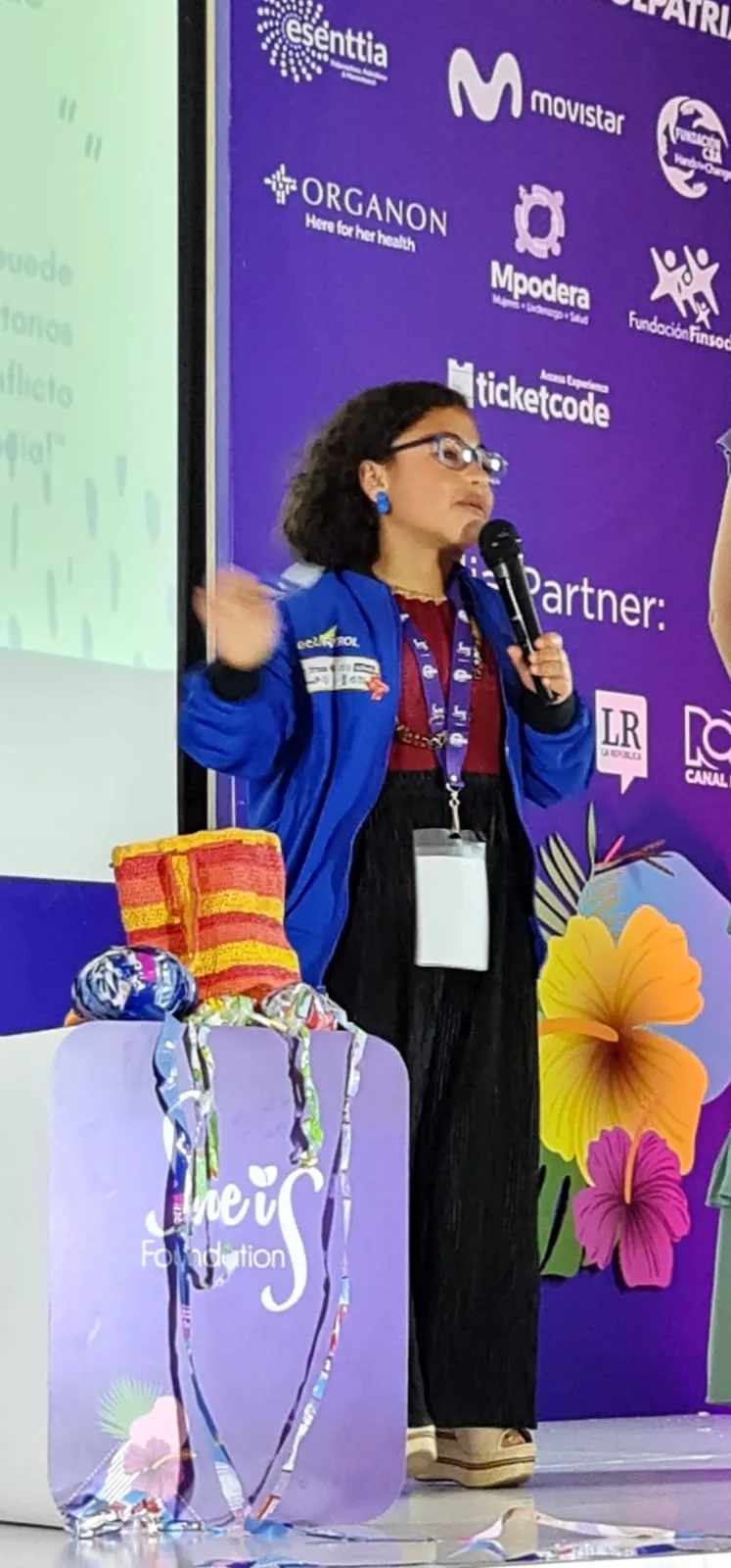 Mujer con chaqueta azul y micrófono habla en el escenario junto a un trofeo de cristal en el She is Global Forum