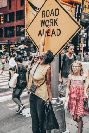 Mujer y una niña de vestido rosa caminan por una calle concurrida junto a una señal «Road work ahead»