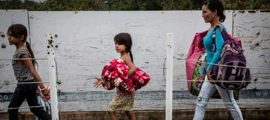 Una mujer y dos niñas caminando con mochilas y bultos junto a un muro blanco
