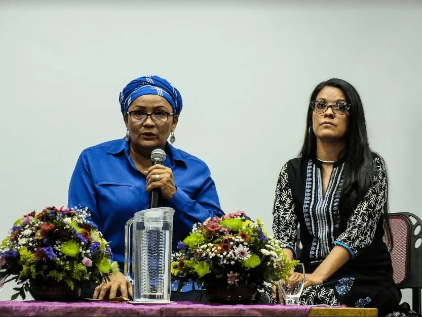 Mujer con pañuelo azul habla al micrófono en un panel, junto a otra mujer y arreglos de flores