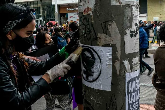 Mujer con tapabocas, guantes y chaqueta de cuero pega un afiche en un poste entre la multitud