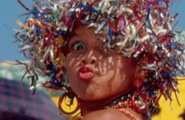 Primer plano de una mujer con peluca de tiras metalizadas de colores que hace un gesto de beso, en un desfile de carnaval